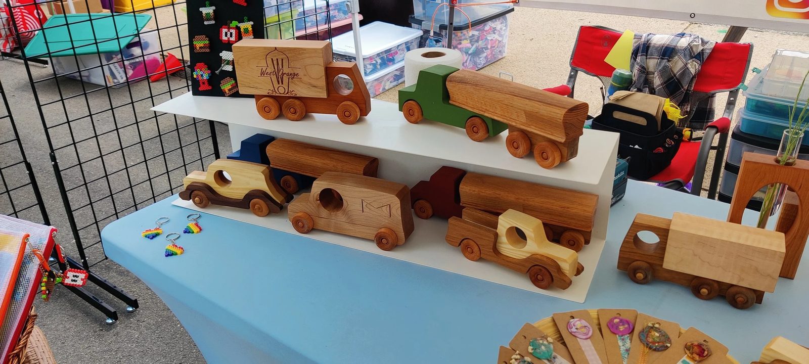 A market-table lineup of handmade wooden toy trucks and cars, including a box truck engraved West Orange