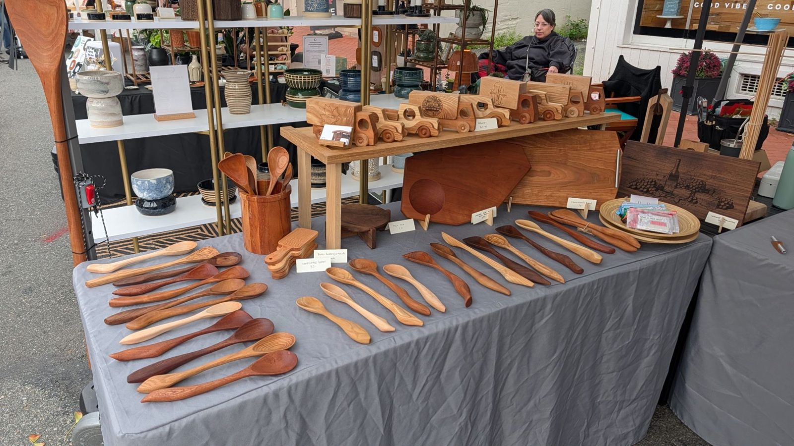 The Max Grossman Designs booth: rows of hand-carved spoons across the table, wooden toy cars and trucks on a riser, and cutting boards behind