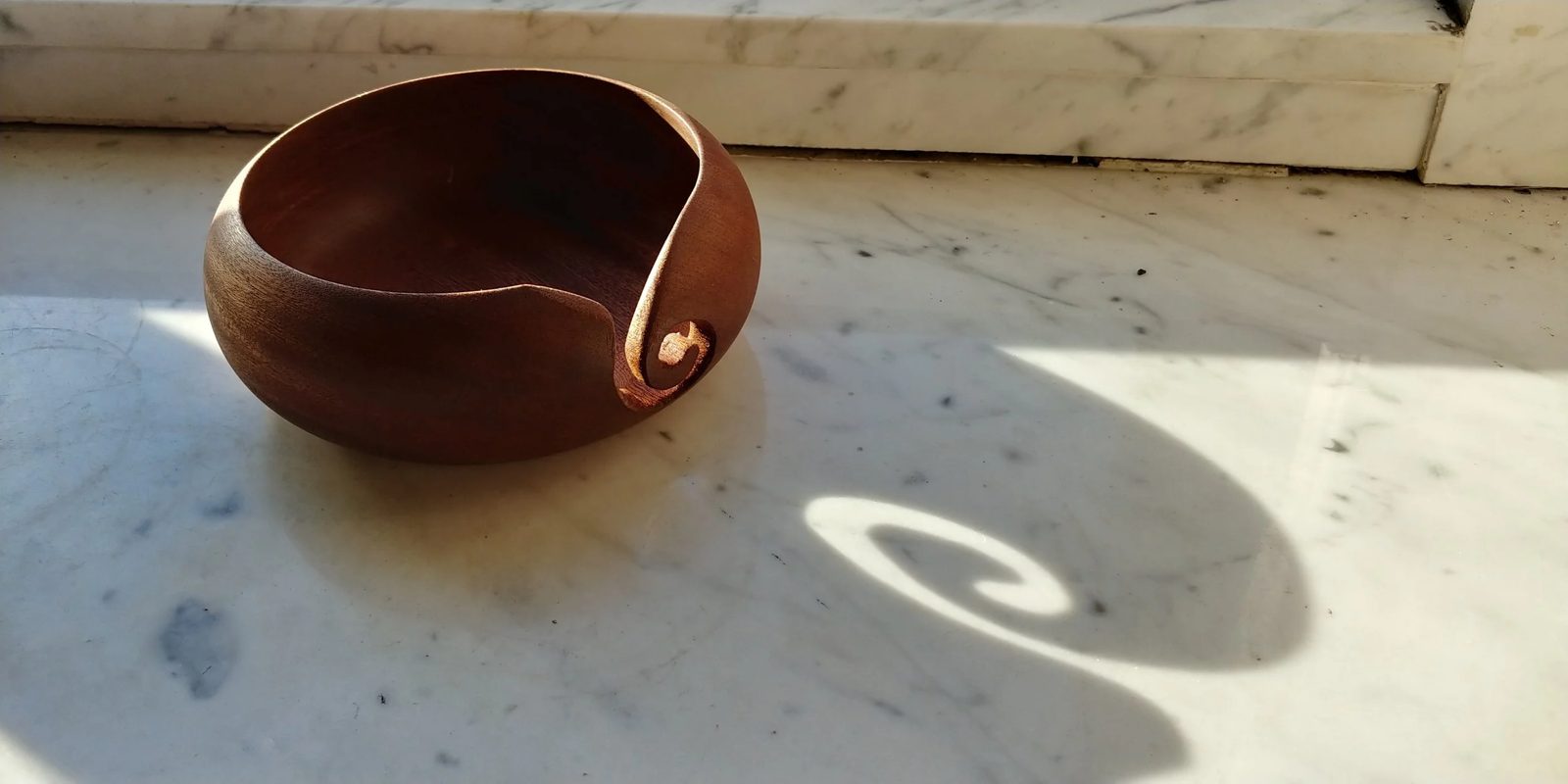 A turned hardwood yarn bowl with a curled slot, on a sunlit marble sill