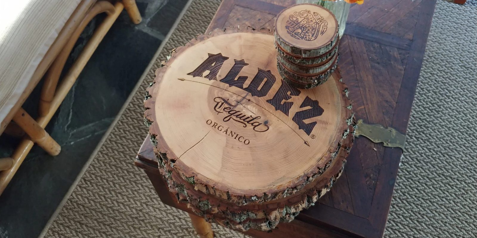 A live-edge serving platter engraved with a tequila brand logo, beside a stack of matching coasters